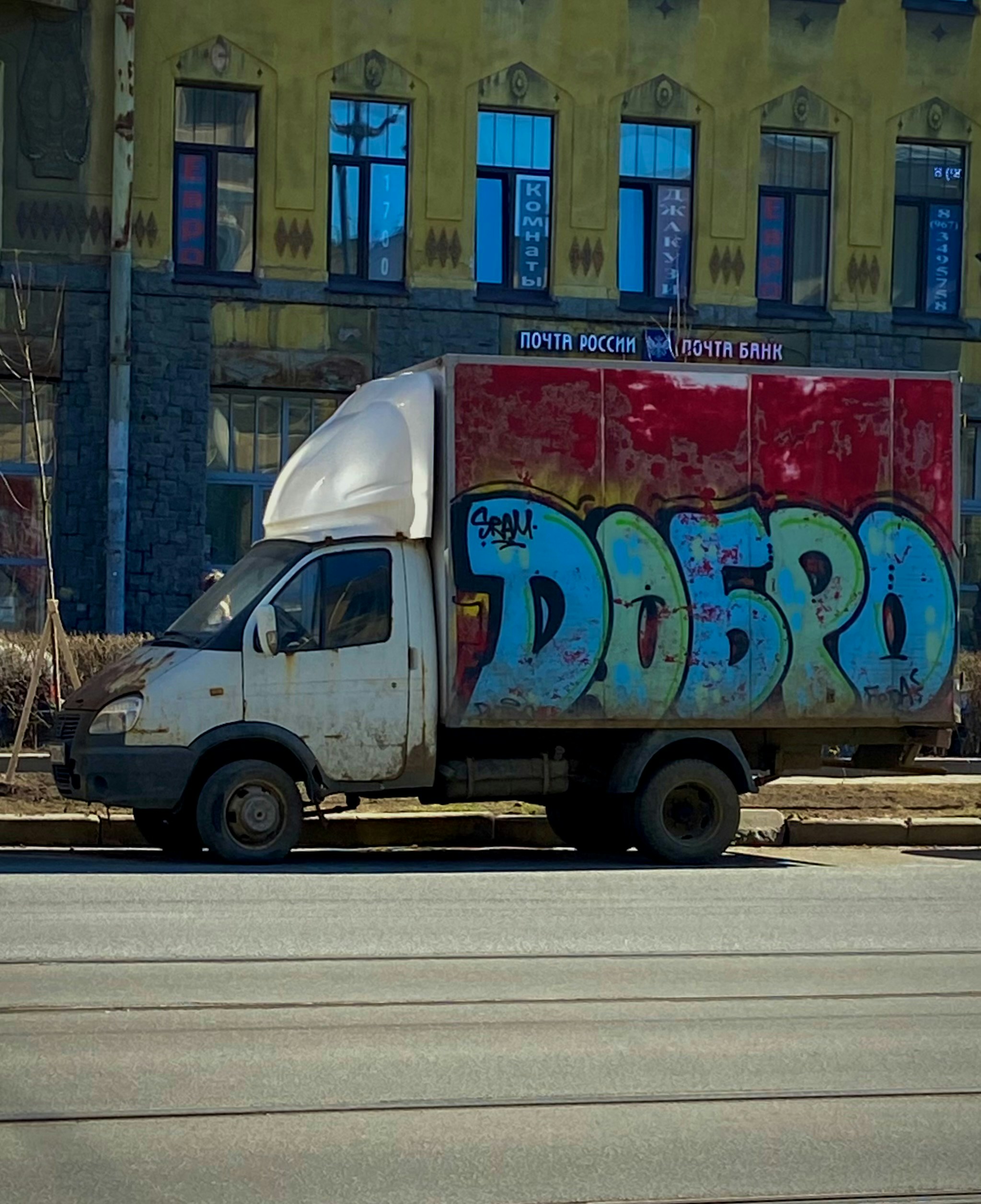 The image features a weathered white GAZelle truck parked in front of a yellow-toned building. Its red cargo panel is covered with bold graffiti spelling “ДОБРО” (Dobro) in stylized blue-green lettering with black outlines. The truck shows signs of wear—scuffs, rust, and faded paint—adding to its urban character. Above the cargo area, a sign reads “ПОЧТА РОССИИ / ПОЧТА БАНК,” while the building’s facade displays decorative moldings and rectangular windows with blue reflections. The scene captures a slice of everyday city life with a touch of street art.
